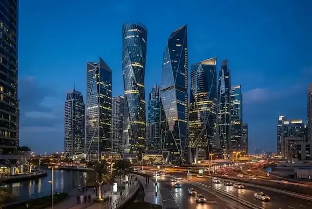 Dusk skyline of angular glass skyscrapers beside a canal, promenade, and busy illuminated roadway.