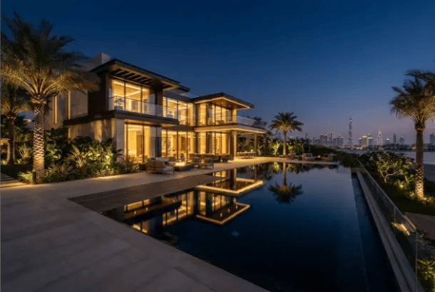 A luxurious modern villa with illuminated glass walls, a glowing infinity pool reflecting the building, and a city skyline view at night.