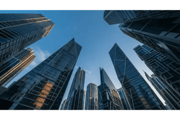 A low-angle worm's-eye view looking straight up at a circle of modern skyscrapers against a clear blue sky.