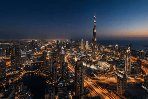 An aerial night view of a bustling city skyline featuring a towering skyscraper illuminated against the dark evening sky, with glowing roads and waterways below.
