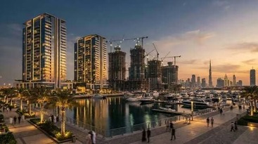 Illuminated marina promenade with yachts, palm trees, high-rises, construction cranes, and a distant skyline at sunset.