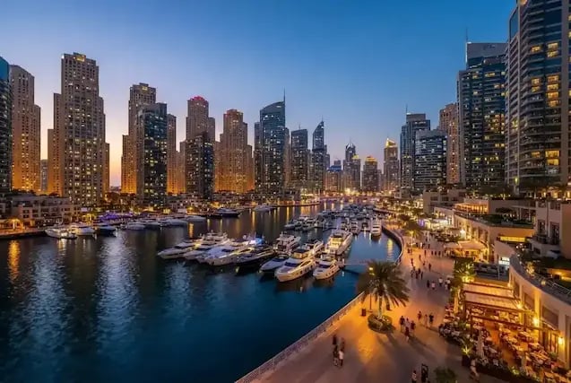 Illuminated high-rise buildings surround a yacht-filled marina and waterfront promenade at dusk.